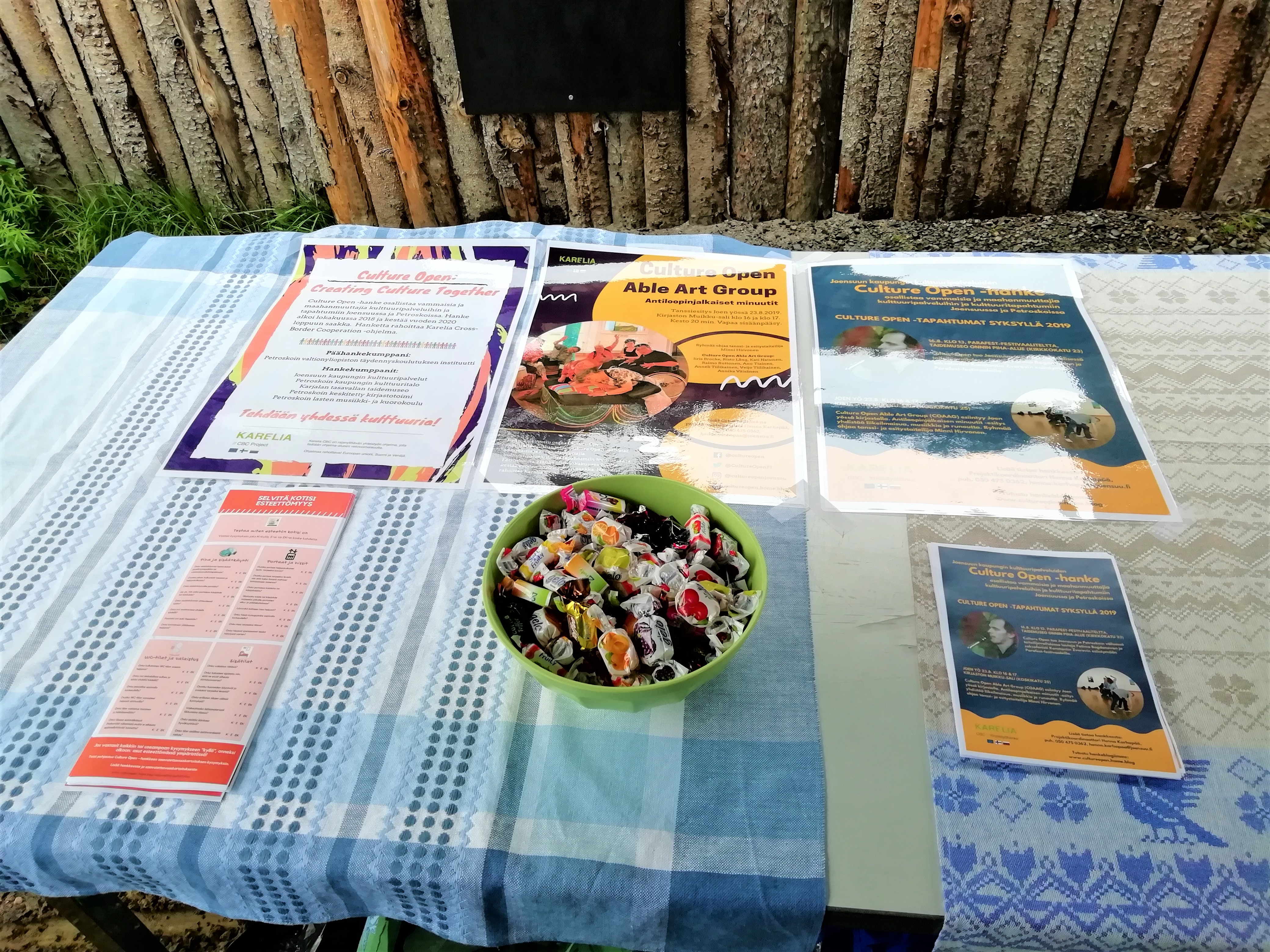 Valokuva, jossa näkyy pöytä. Pöydälle on aseteltu hankejuliste, paperisia esteettömyystestejä sekä tapahtumaesite syksylle 2019.